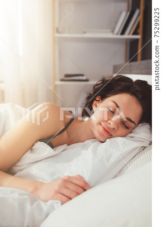 Woman lying in bed and keeping eyes closed while covered with blanket Woman lying in bed and keeping eyes closed while covered with blanket 75299225