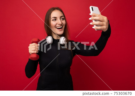 Attractive charming young smiling happy woman holding and using mobile phone taking selfie wearing 75303670