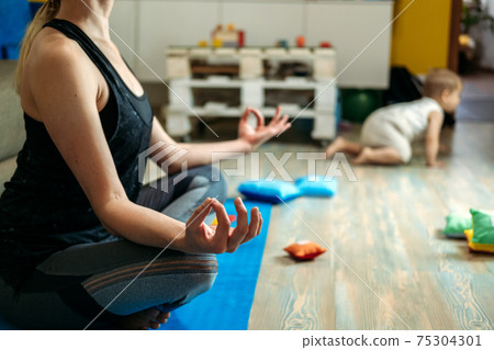 Mindfulness meditation concept. Young woman, mother practicing yoga at home sitting in lotus pose, meditating relaxing with closed eyes with baby daughter at home. Mindfulness meditation concept Mindfulness meditation concept. Young woman, mother practicing yoga at home sitting in lotus pose, meditating relaxing with closed eyes with baby daughter at home. Mindfulness meditation concept 75304301