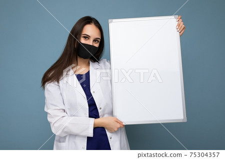 Young nurse in protective face mask and white medical coat holding an empty magnetic board isolated Young nurse in protective face mask and white medical coat holding an empty magnetic board isolated 75304357