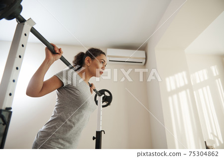 Beautiful woman training with a barbell at home gym. Active and healthy lifestyle Beautiful woman training with a barbell at home gym. Active and healthy lifestyle 75304862