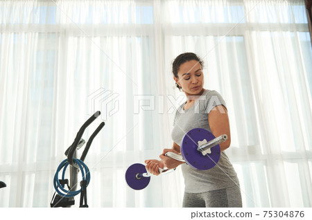 Determined sporty woman looking at her biceps muscle while pumping hands during heavy barbell workout 75304876