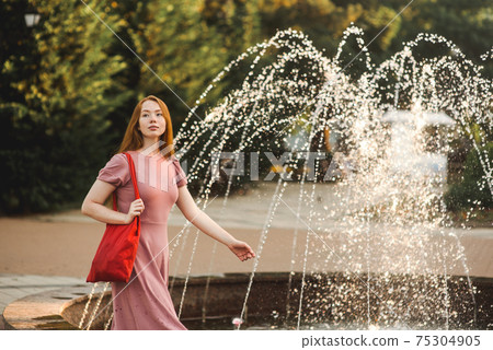 Young beautiful red-haired long-haired woman in a summer pink dress walks through the streets of a summer city. 75304905