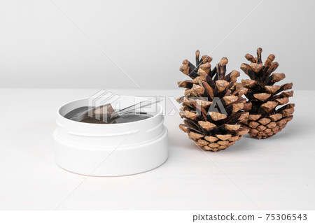A white box with golden eye patches on the white table with pine cones. Cosmetics product for skin care A white box with golden eye patches on the white table with pine cones. Cosmetics product for skin care 75306543