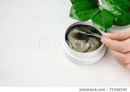 Close up woman hand taking a golden eye patch with spatula from the white box with green leaf on the table with copy space. Close up woman hand taking a golden eye patch with spatula from the white box with green leaf on the table with copy space. 75306544