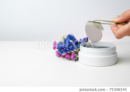 Close up woman hand holding a golden eye patch with tweezers on the white background and colorful flowers with copy space. Close up woman hand holding a golden eye patch with tweezers on the white background and colorful flowers with copy space. 75306545