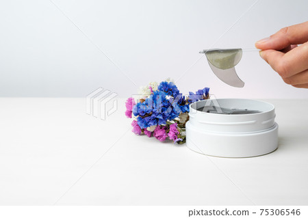 Close up woman hand holding a golden eye patch with spatula on the white background and colorful flowers with copy space. Close up woman hand holding a golden eye patch with spatula on the white background and colorful flowers with copy space. 75306546