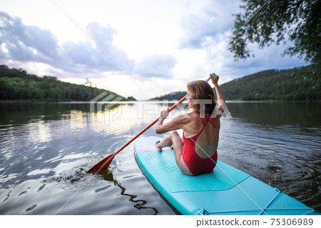 Rear view of senior woman paddleboarding on lake in summer. Rear view of senior woman paddleboarding on lake in summer. 75306989