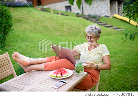 Happy senior woman with laptop working outdoors in garden, home office concept. 75307013