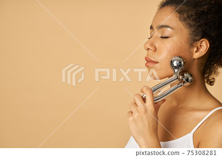 Relaxed young woman posing with eyes closed while using silver metal face roller isolated over beige background 75308281