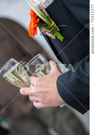 The groom holds a glass or flute of champagne on the background of the friends on nature at the The groom holds a glass or flute of champagne on the background of the friends on nature at the 75311777