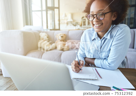 Happy mixed race teen girl in glasses smiling while doing homework using laptop, studying in the living room at home during quarantine 75312392