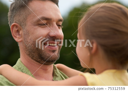 Close up of cheerful dad looking at his little girl, holding her while spending time together outdoors on a warm day Close up of cheerful dad looking at his little girl, holding her while spending time together outdoors on a warm day 75312559