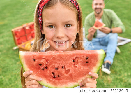 Close up portrait of lovely little girl looking at camera while holding watermelon slice, having a picnic with her daddy in the park Close up portrait of lovely little girl looking at camera while holding watermelon slice, having a picnic with her daddy in the park 75312645