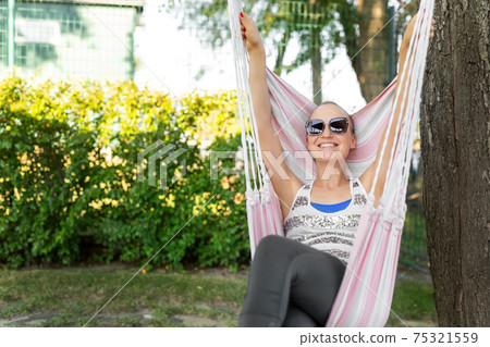 Young adult happy short haired shaved bald attractive woman chilling enjoy relaxing sitting in fabric chair hammock at yard in garden near house. Peaceful and idyllic outdoor rest lifestyle 75321559