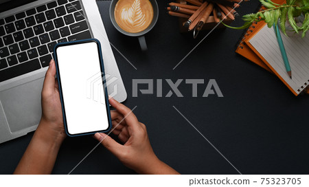 Overhead shot of woman hands holding mobile phone with empty screen on modern dark workplace. Overhead shot of woman hands holding mobile phone with empty screen on modern dark workplace. 75323705