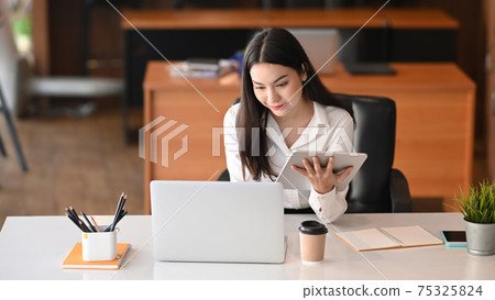Smiling businesswoman holding digital tablet and working on laptop computer at office desk. Smiling businesswoman holding digital tablet and working on laptop computer at office desk. 75325824