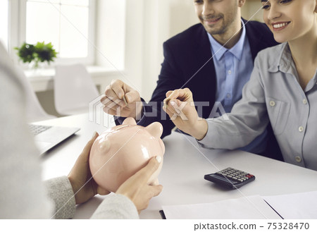 Couple puts coin in piggy bank sitting at desk with bank manager or insurance broker 75328470