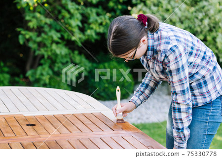 young girl painting wooden exotic wood table in the garden with a brush - shallow depth of field 75330778