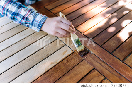 hand holding a brush applying varnish paint on a wooden garden table - painting and caring for wood with oil 75330781
