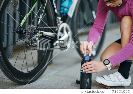 Cropped shot of young woman, professional female cyclist taking pump for inflating the tire of her bicycle, kneeling outdoors on a daytime Cropped shot of young woman, professional female cyclist taking pump for inflating the tire of her bicycle, kneeling outdoors on a daytime 75331095