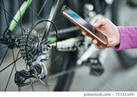 Close up shot of hand of female cyclist holding smartphone while checking her bicycle mechanisms, sprocket and chain on a mountain bike outdoors on a daytime 75331104