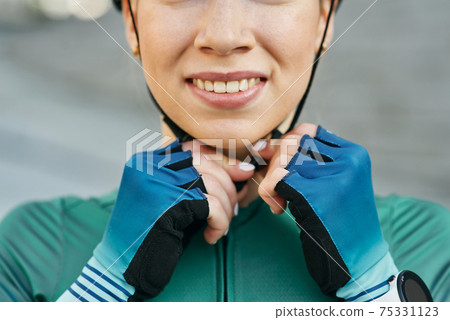Cropped portrait of young woman, female cyclist smiling and fastening her cycling helmet while posing outdoors on a daytime 75331123