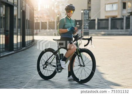 Professional female cyclist in cycling garment and protective gear looking at camera, holding smartphone while riding bicycle in city center. Sportswoman training, exercising outdoors Professional female cyclist in cycling garment and protective gear looking at camera, holding smartphone while riding bicycle in city center. Sportswoman training, exercising outdoors 75331132