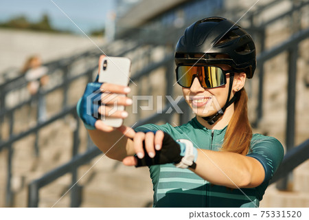 Portrait of cheerful professional female cyclist in cycling garment and protective gear smiling while taking selfie using smartphone, resting after riding bicycle in city center 75331520