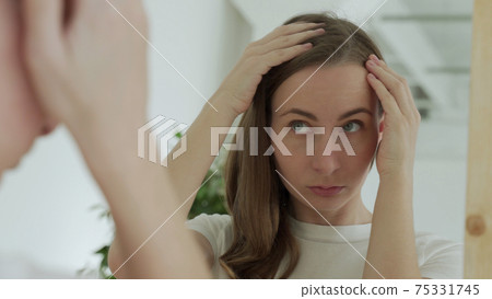 Sad upset woman standing by mirror looking at matted fragile strand in her hand thinking about natural medicinal shampoo restoring hair structure Sad upset woman standing by mirror looking at matted fragile strand in her hand thinking about natural medicinal shampoo restoring hair structure 75331745