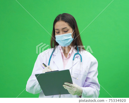 Young Asian woman doctor taking notes, making a report on paper notepad with a face mask and doctor gown uniform coat and stethoscope isolated on green background, medical treatment. Coronavirus. Young Asian woman doctor taking notes, making a report on paper notepad with a face mask and doctor gown uniform coat and stethoscope isolated on green background, medical treatment. Coronavirus. 75334920