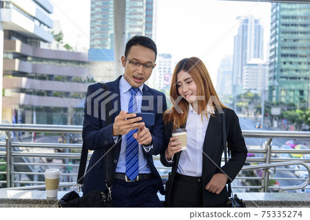 Businessman Businesswoman drinking coffee in town using smartphone outside office modern city. Hands holding take away coffee cup and smart phone talking together Business partner with cup of coffee Businessman Businesswoman drinking coffee in town using smartphone outside office modern city. Hands holding take away coffee cup and smart phone talking together Business partner with cup of coffee 75335274