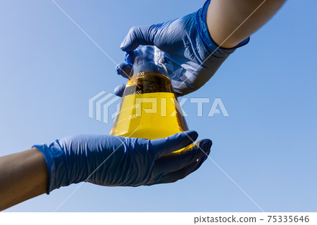 Hand with gloves holding beaker with ethanol biofuel against blue sky 75335646