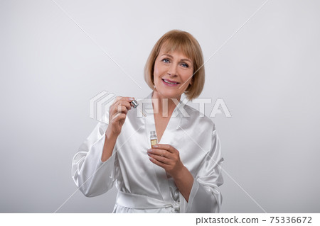 Cheerful senior lady in dressing gown using face serum on light studio background. Skincare and wellness Cheerful senior lady in dressing gown using face serum on light studio background. Skincare and wellness 75336672