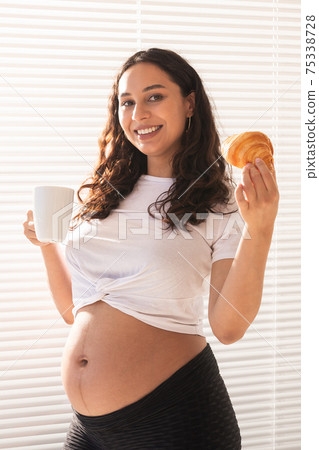 Beautiful pregnant woman holding croissant and cup of coffee in her hands during morning breakfast. Concept of good health and positive attitude while expecting baby 75338728