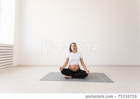 Healthy beautiful pregnant woman meditates before yoga class and relaxes while sitting on mat on the floor. Concept of physical and mental preparation of body for childbirth. Copyspace 75338729