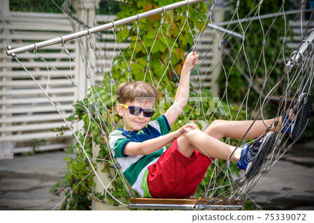 Little adorable kid sitting on chain swing during summer holidays on vacation. Boy relaxed and happy wearing casual clothes in sunglasses. Active summer leisure for kids in the city concept Little adorable kid sitting on chain swing during summer holidays on vacation. Boy relaxed and happy wearing casual clothes in sunglasses. Active summer leisure for kids in the city concept 75339072