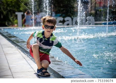 Little boy plays in the square near pool with water jets in the fountain at sunny summer day. Active summer leisure for kids in the city Little boy plays in the square near pool with water jets in the fountain at sunny summer day. Active summer leisure for kids in the city 75339074