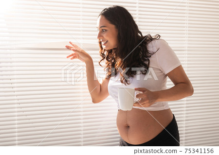 Curious young beautiful pregnant woman drinking tea and looking through the blinds at the window. Concept of joy and good news while waiting for baby Curious young beautiful pregnant woman drinking tea and looking through the blinds at the window. Concept of joy and good news while waiting for baby 75341156