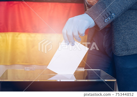 Side view close-up of man's hand with protective glove putting vote in ballot box against Germany flag background. Side view close-up of man's hand with protective glove putting vote in ballot box against Germany flag background. 75344852