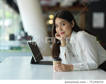 Side view of businesswoman smiling with hand to cheek and holding paper cup in workplace 75349434