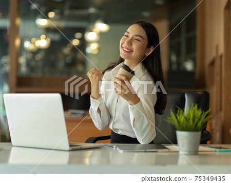 Female office worker celebrating on her achievement while holding paper cup and sitting at workplace 75349435