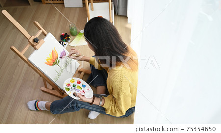 Over head shot of a young female artist in yellow sweater with palette in her hand is painting on canvas at studio. 75354650