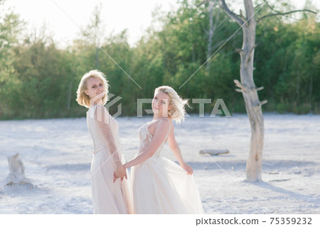 Beautiful lesbian couple walking on sand along river bank on their wedding day Beautiful lesbian couple walking on sand along river bank on their wedding day 75359232