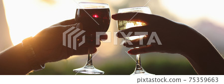 Male and female hands are holding glass of red and white wine against sunset background. 75359663