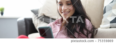 Woman sits on floor next to sofa and looks smiling at smartphone screen. 75359869