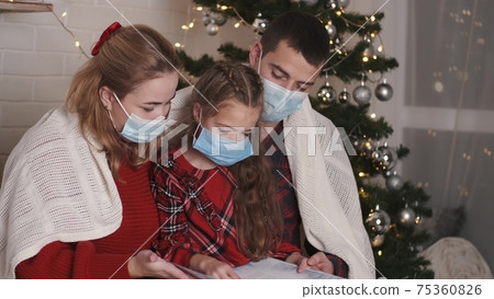 Happy Caucasian family in masks sitting at decorated home near Christmas glowing tree on New Year's Eve together Happy Caucasian family in masks sitting at decorated home near Christmas glowing tree on New Year's Eve together 75360826