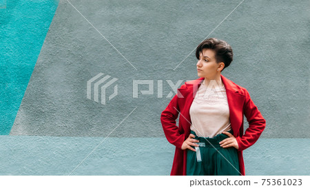 Candid portrait Young Brunette woman with short hair on blue wall background. Outdoor portrait modern gen z woman in red coat 75361023