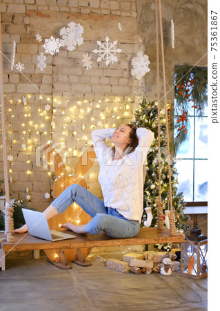 Female student sitting on swing with laptop in decorated room by garlands and Christmas tree. 75361867
