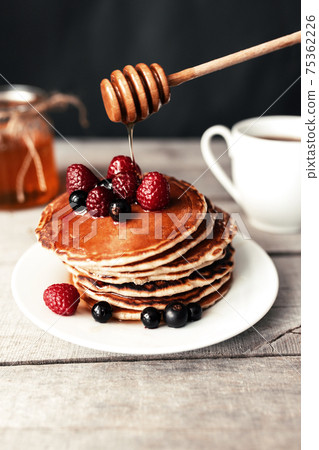 Pancakes with berries and honey on a white plate, wooden spoon, jar, coffee cup Pancakes with berries and honey on a white plate, wooden spoon, jar, coffee cup 75362226